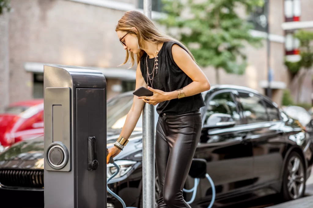 Young Woman Charging Electric Car Standing With Smart Phone Outdoors Street